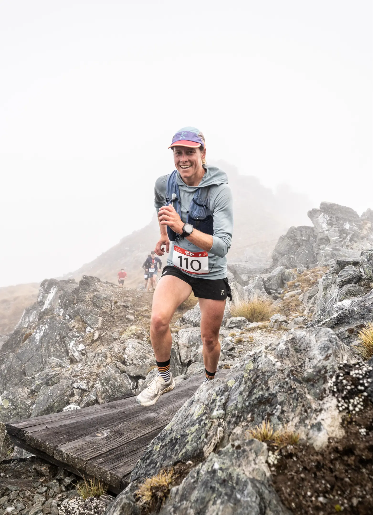 A trail runner jogs across a plank of wood on a rocky mountain trail.