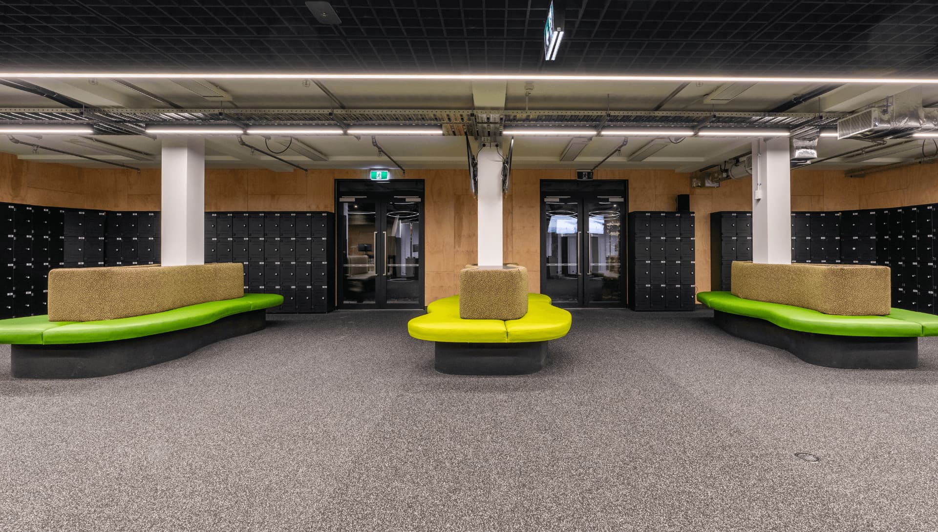 A modern locker room with black lockers lining the walls, green and tan curved bench seating in the center, and industrial-style ceilings with exposed piping and lighting.