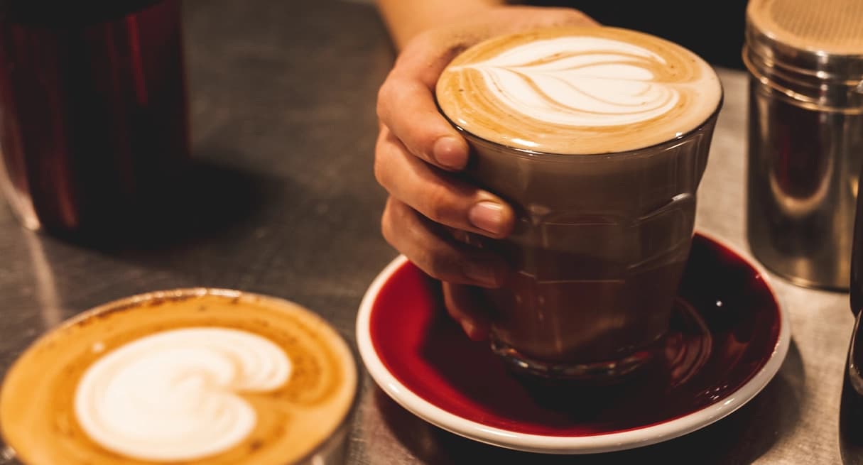 Two lattes with latte art served in glass cups on red saucers, one being held by a hand.