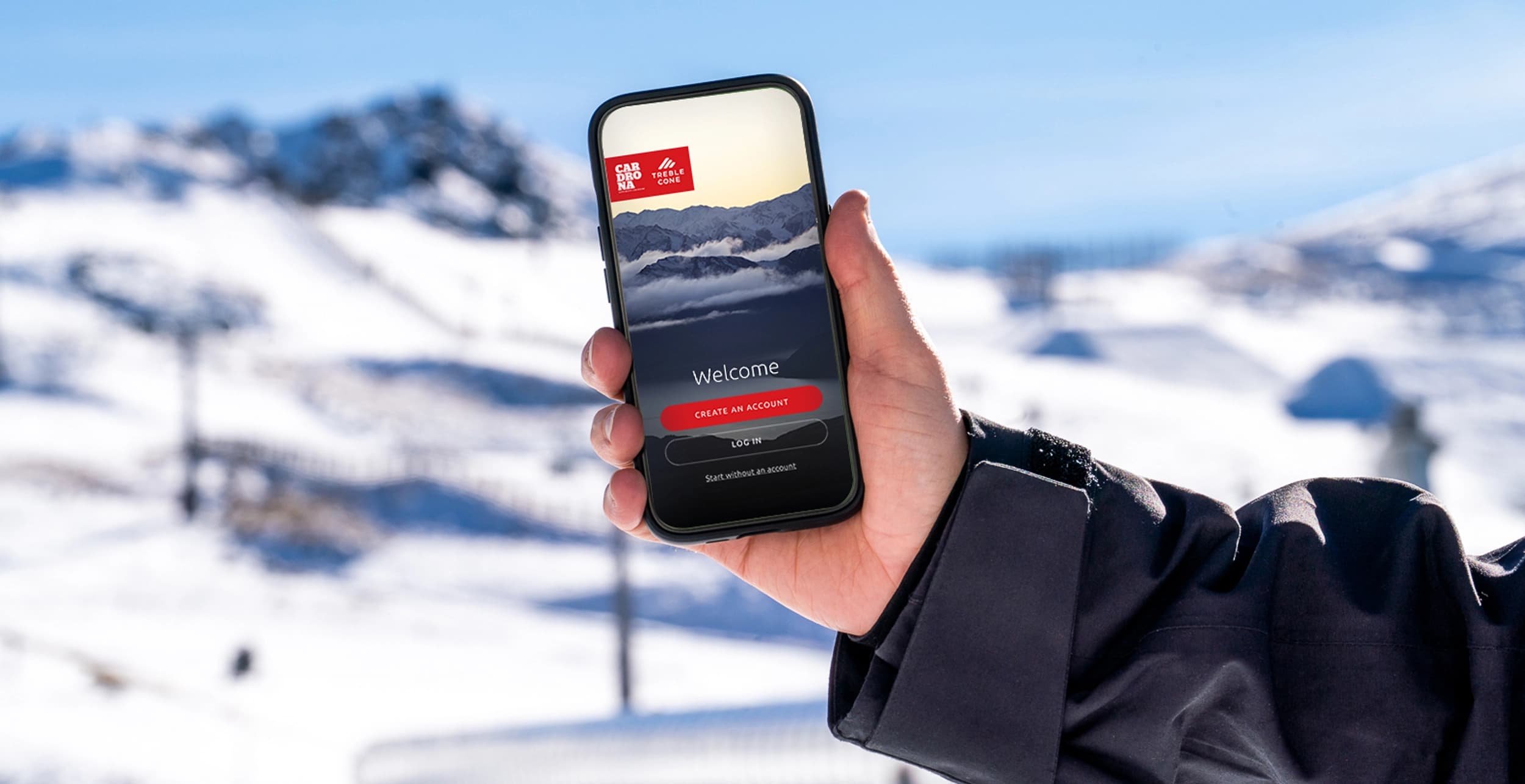 A hand holding a smartphone displaying the Cardrona and Treble Cone ski resort app welcome screen, with a snowy mountain landscape in the background.