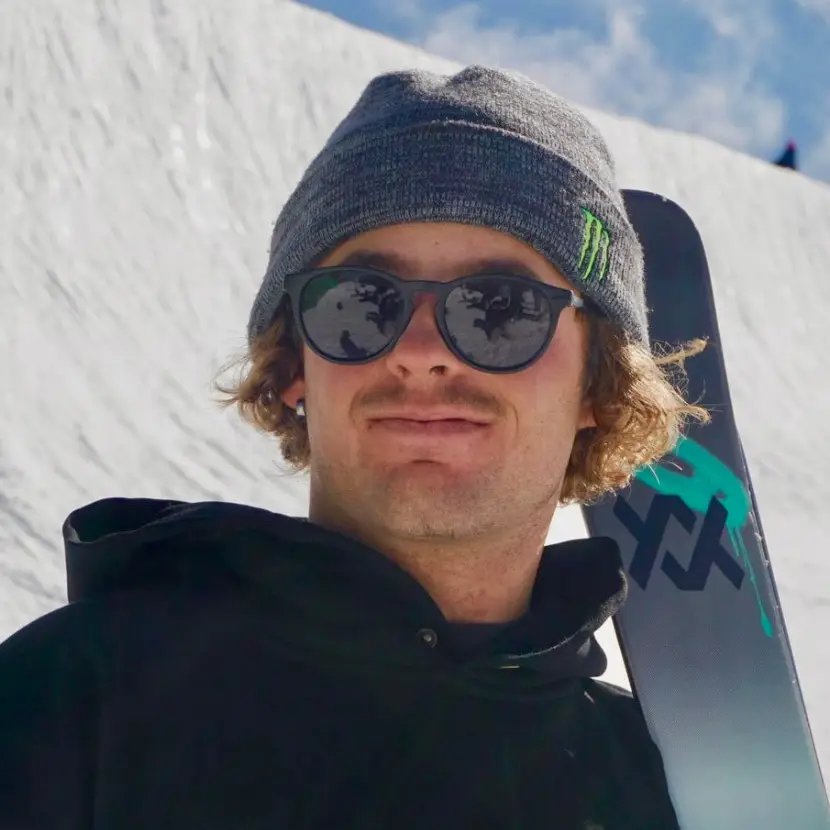 Close up headshot of a skier wearing a Monster Energy branded beanie and sunglasses with a snowy slope in the background.