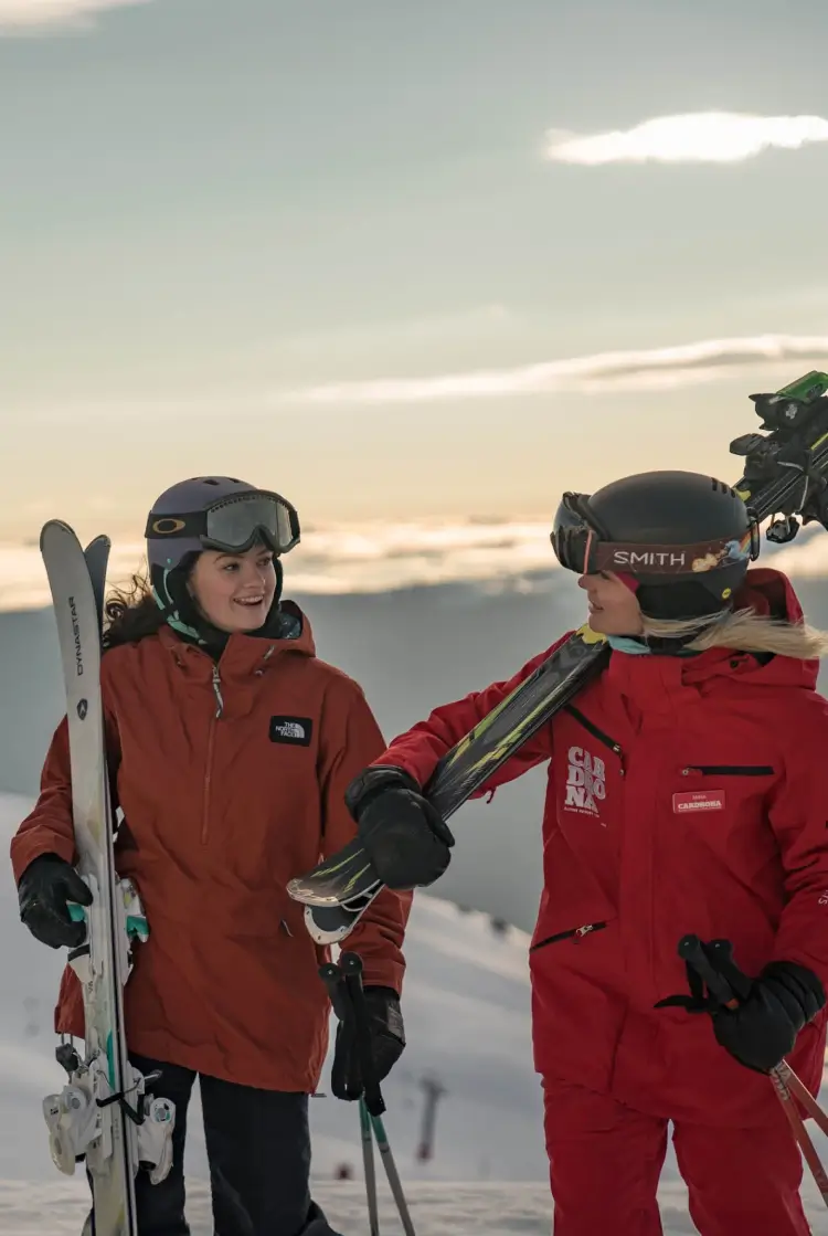 An instructor and a learner dressed in winter gear talk at Cardrona Alpine Resort, holding skis with a scenic mountain view in the background at sunset.