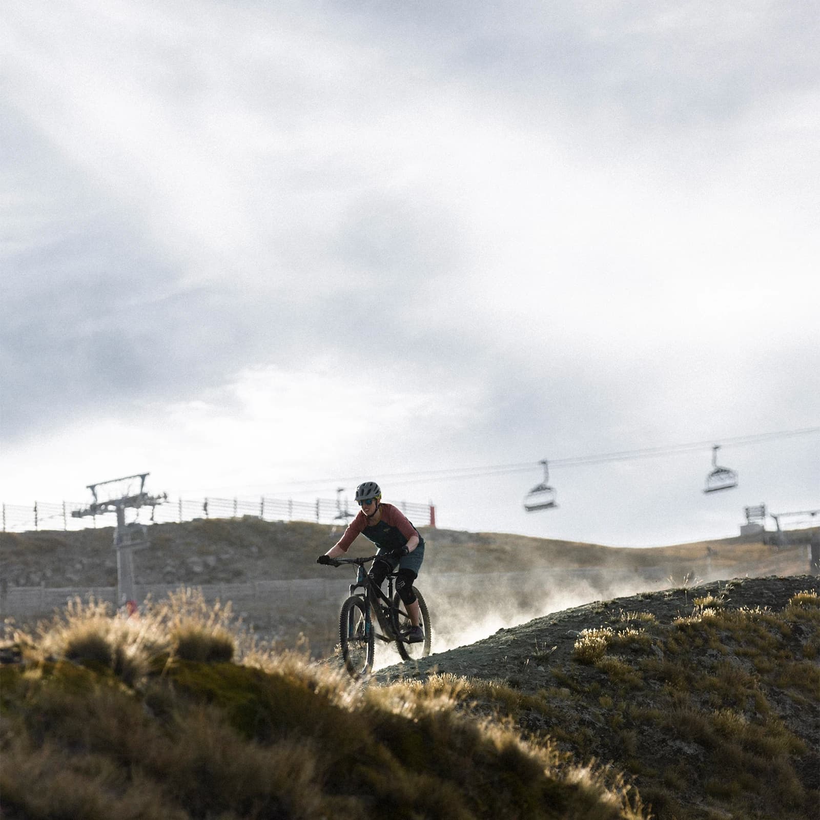 A mountain biker, dressed in a red and black outfit and wearing a helmet, rides along a dusty trail. In the background, a chairlift with several gondolas moves over the rugged mountainous landscape under an overcast sky.