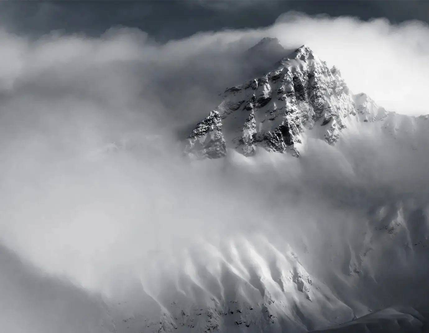 A towering, snow-covered mountain peak partially shrouded in swirling clouds, creating a dramatic and mysterious atmosphere.