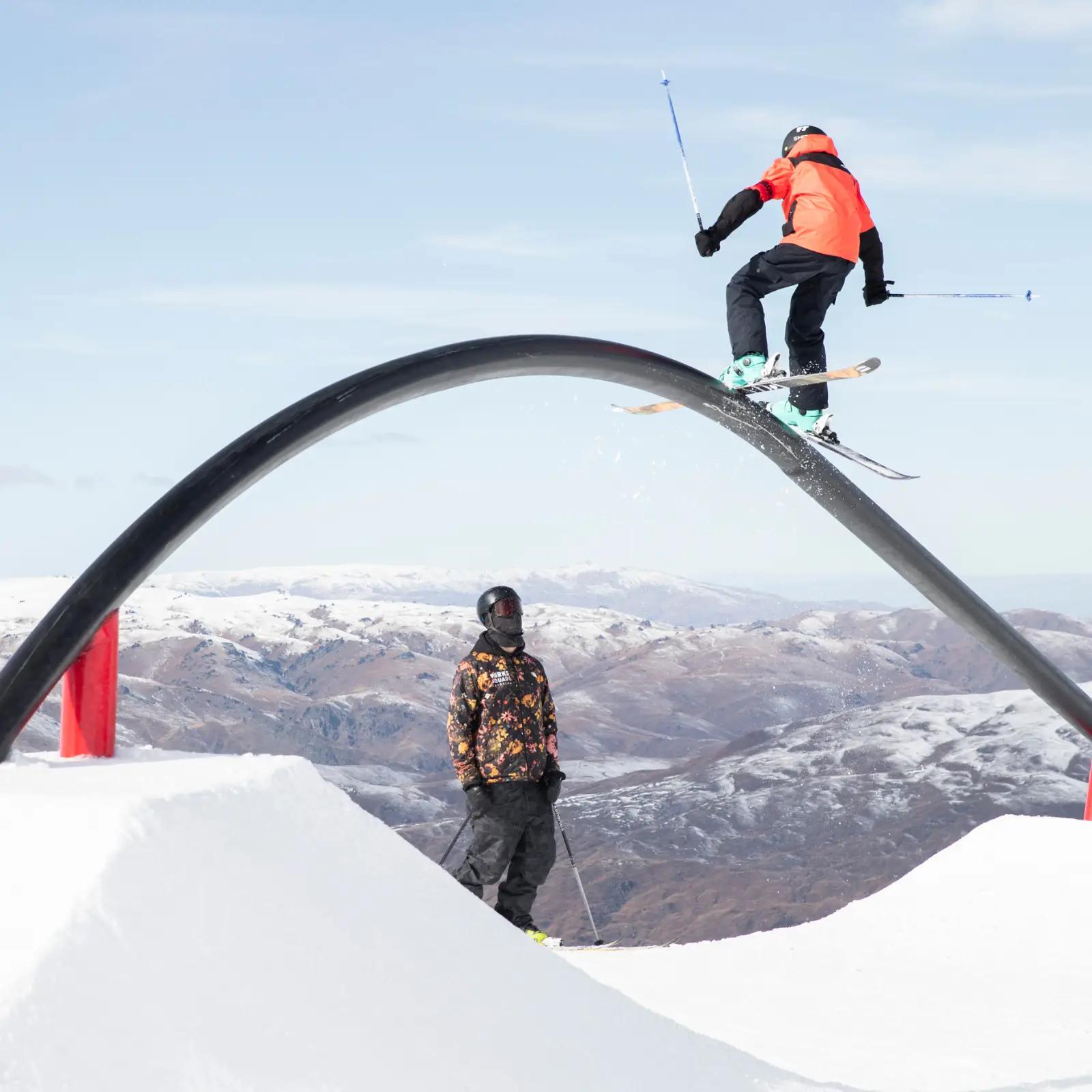 A skier in an orange jacket grinds a curved rail feature in a snow park, with a coach or fellow athlete watching from below. Snow-covered mountains stretch across the background under a clear blue sky.