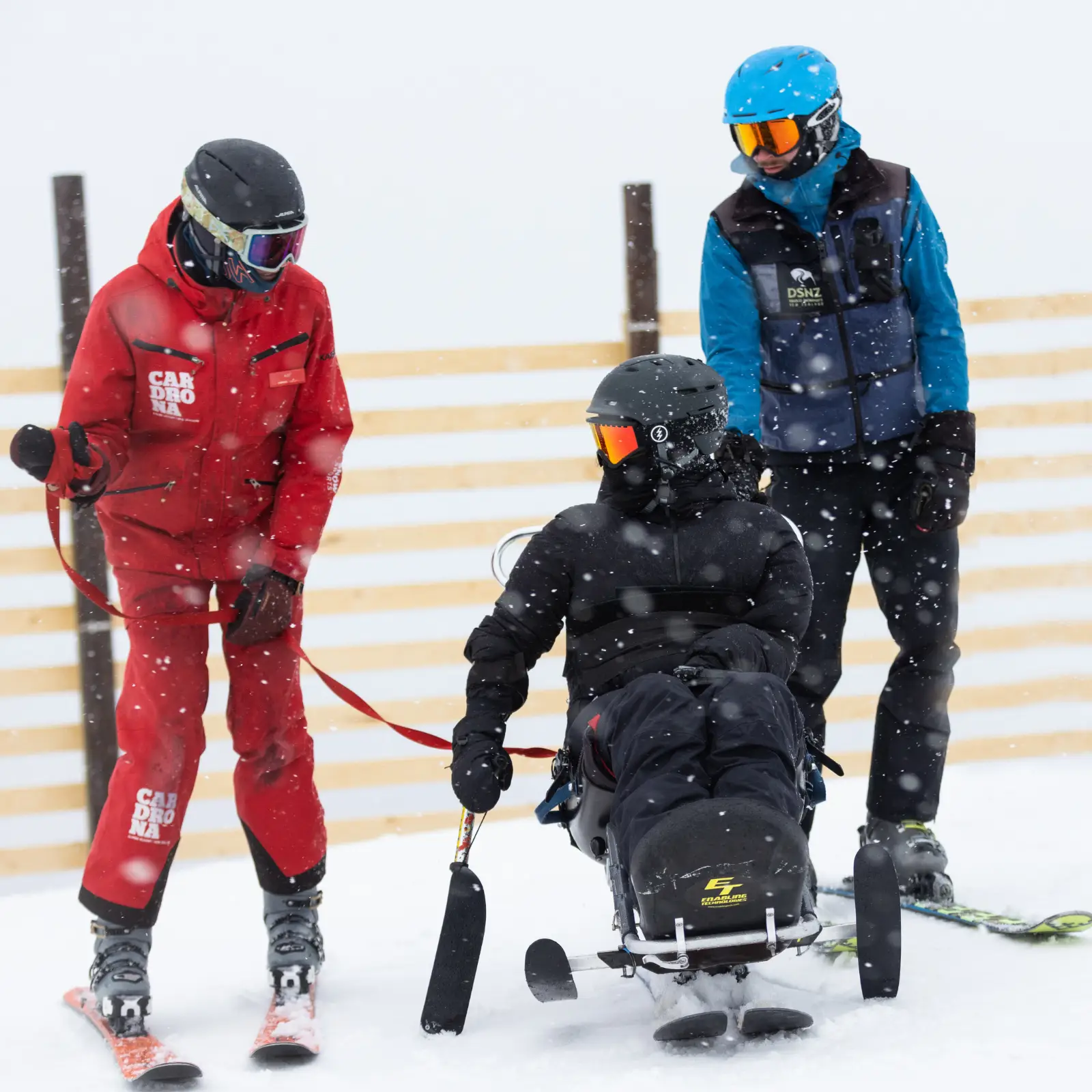 Two skiers, one in Cardrona-branded red ski clothes, assist a sit-skier on a snowy day.