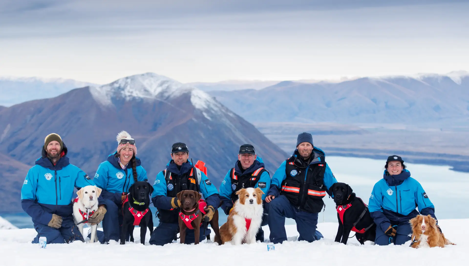 Ski patrol team and their dogs pose on the top of a snowy mountain with a lake view in the background.