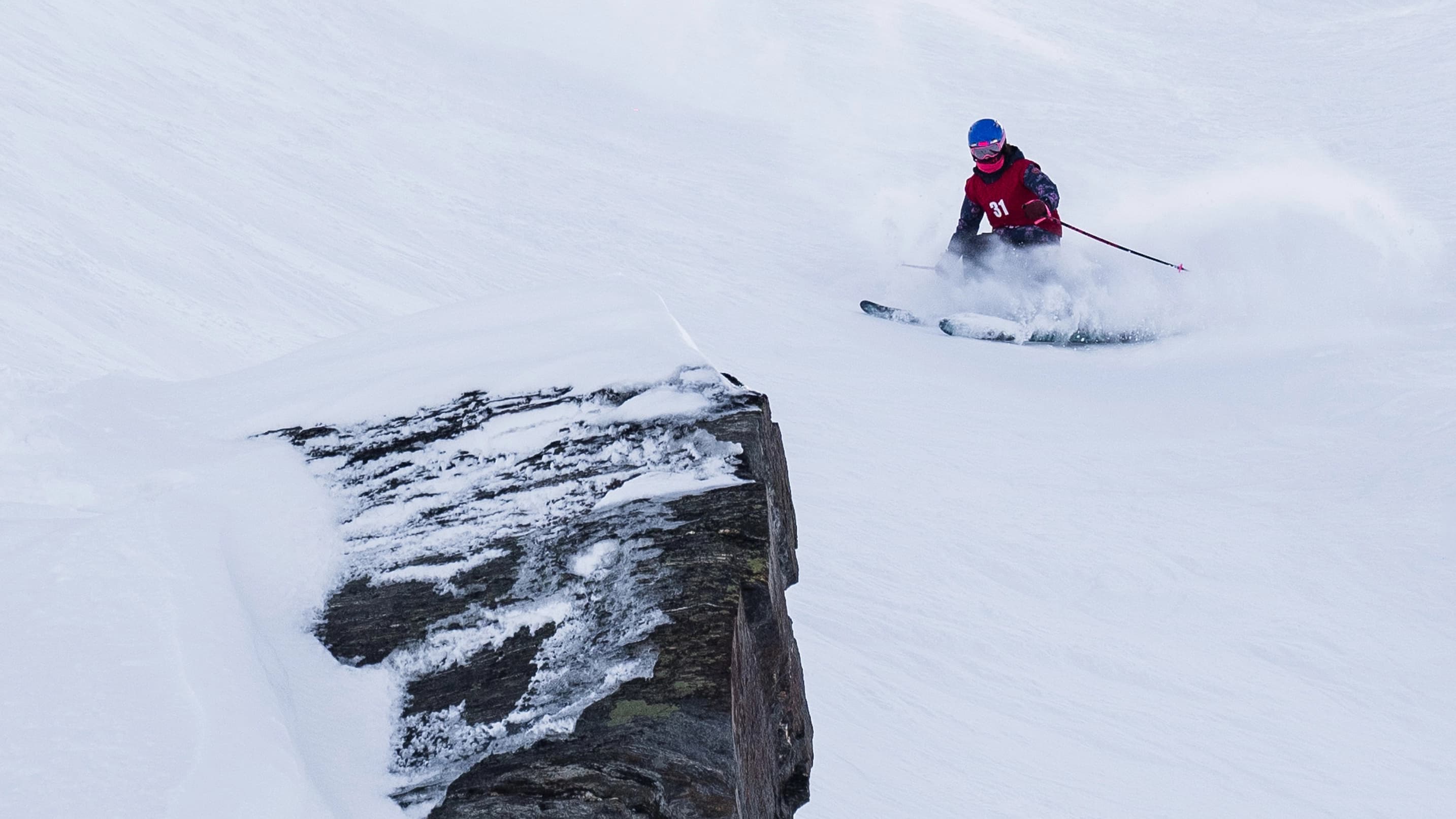 Young skier carving fresh snow near a rock face in a junior freeride competition.
