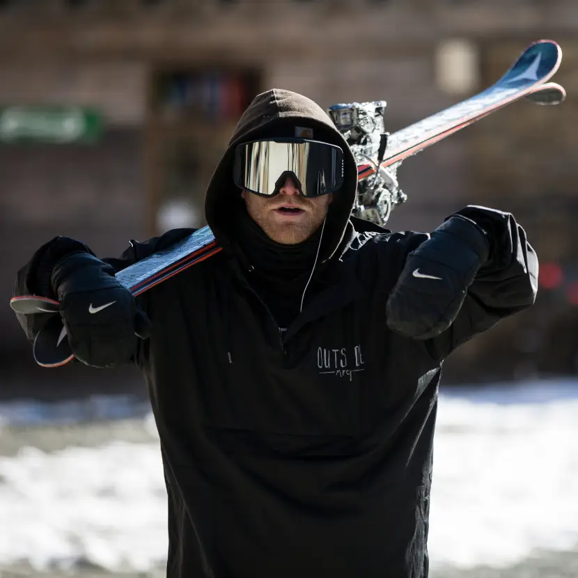 A skier wearing a black hoodie, black gloves and goggles walks with his skies over his shoulder on snowy ground.