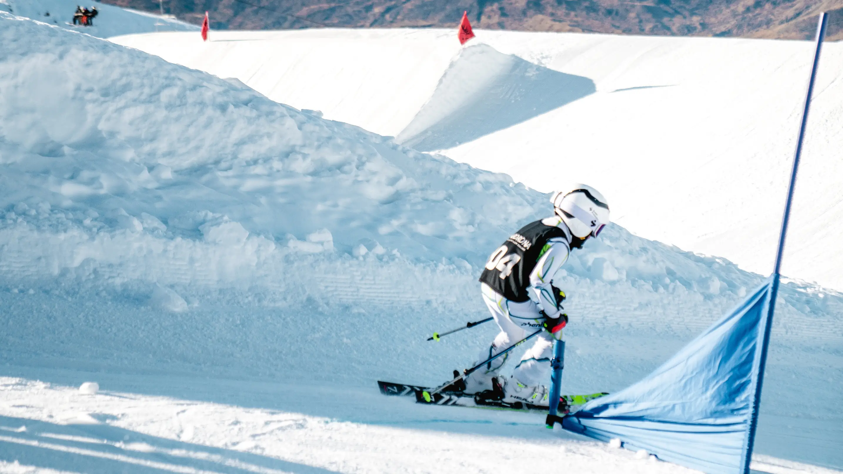 Young skier in full gear races through a blue slalom gate on a snowy mountain course, with rugged alpine peaks and terrain park features in the background.