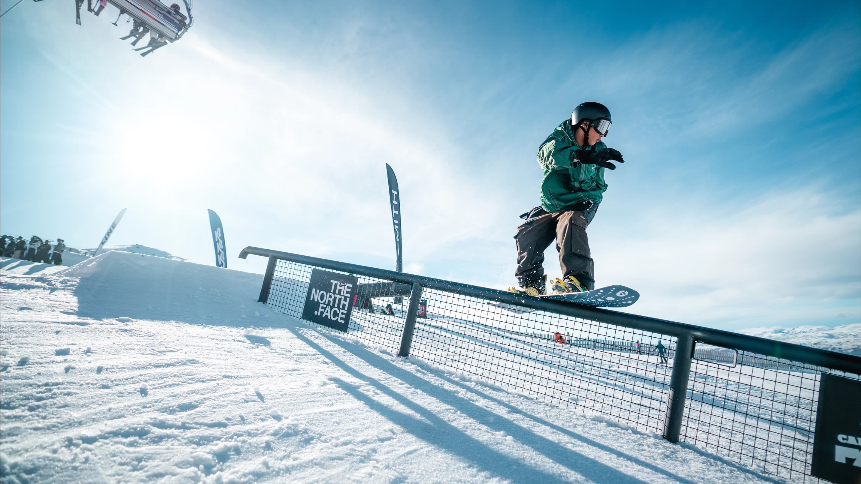 A snowboarder rides a North Face rail feature with flags and blue skies behind.