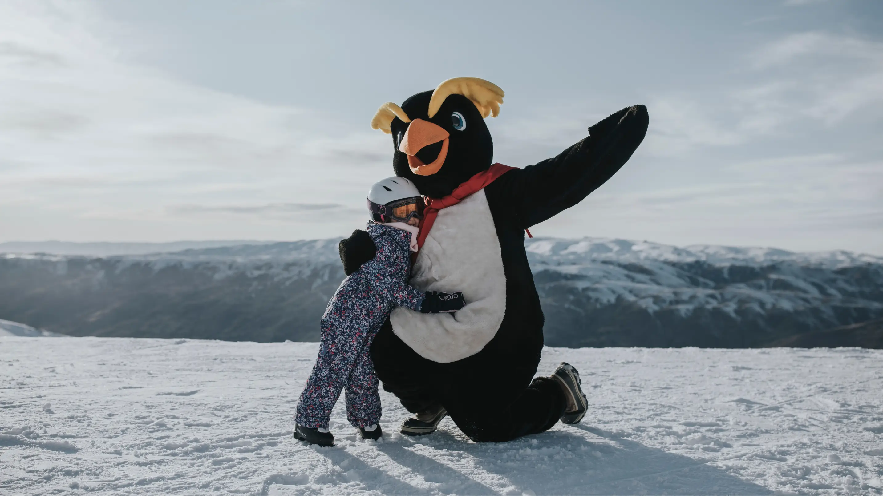 A penguin mascot poses hugging a child, dressed in full ski gear, on a snowy mountain.