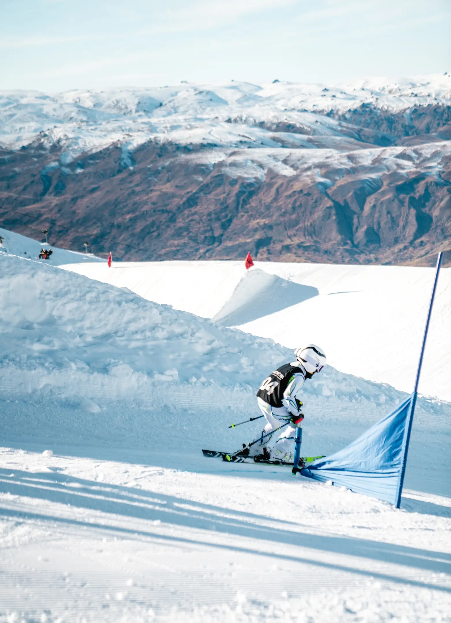 Young skier in full gear races through a blue slalom gate on a snowy mountain course, with rugged alpine peaks and terrain park features in the background.