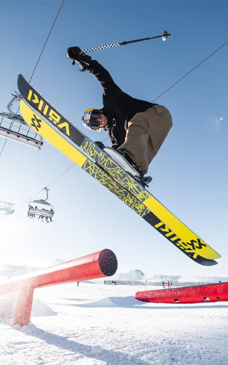 A skier performs an aerial trick under a clear blue sky, with a ski lift and mountain backdrop visible in the background.