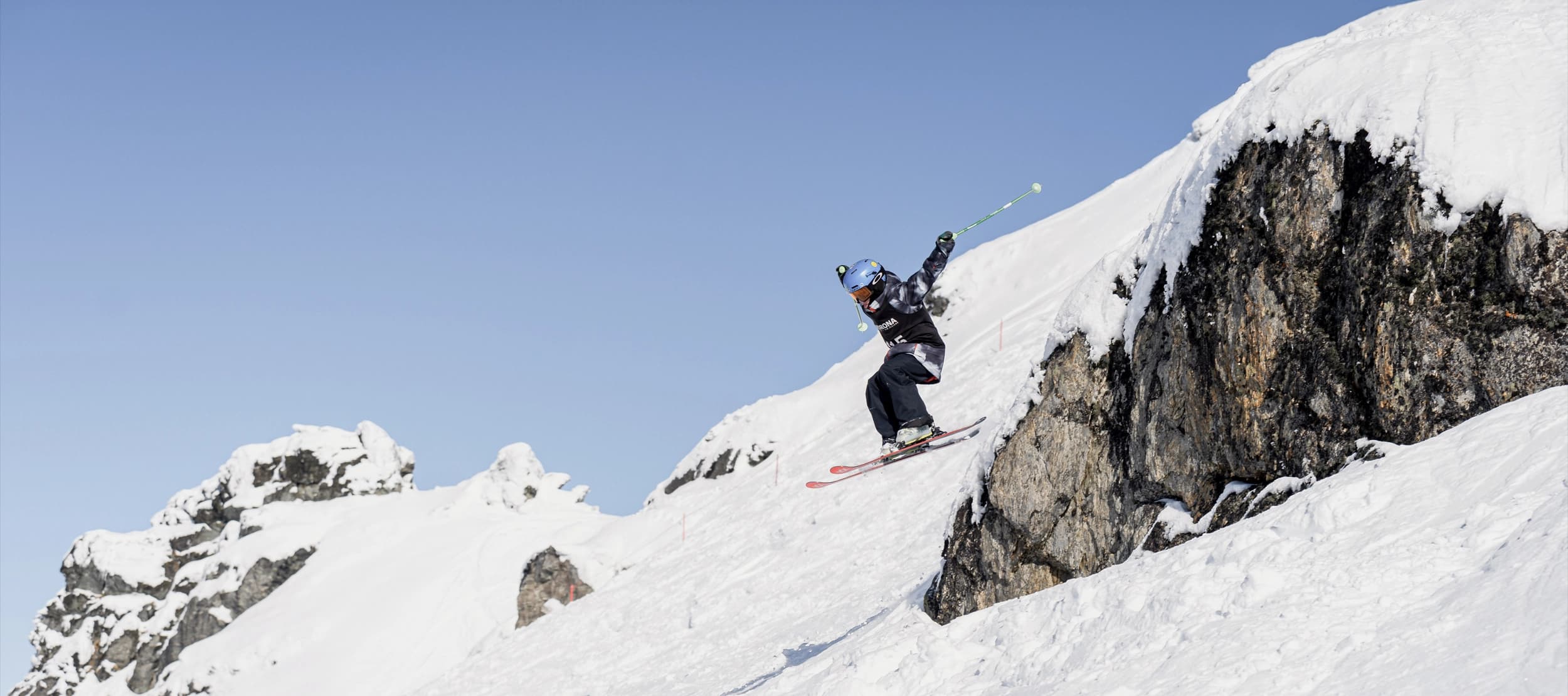 A skier in mid-air launches off a cliff edge against a bright blue sky.