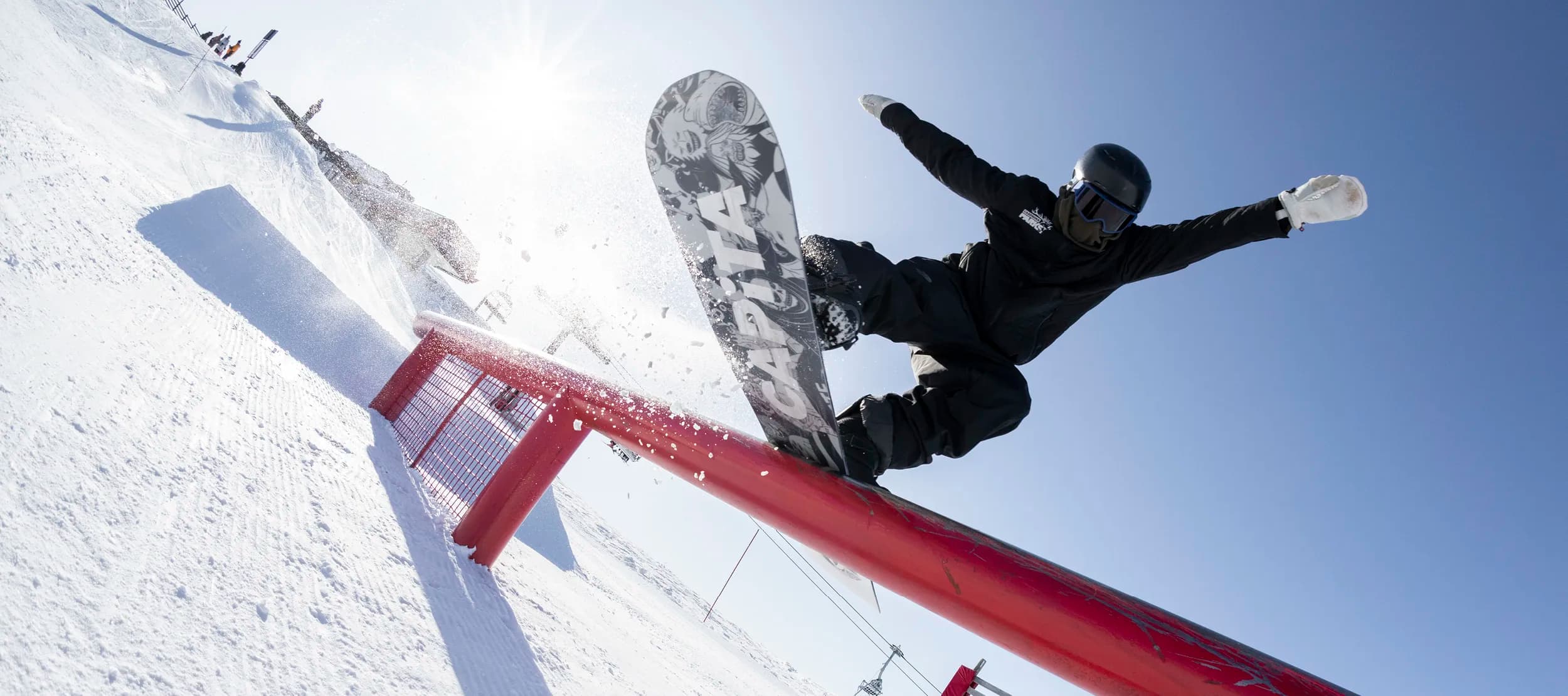 A snowboarder performing a trick while airborne over a bright red rail in a snow-covered terrain park. The rider is in mid-motion, board vertical to the ground, showcasing the artwork on the underside of the board. There is a snowy mountain landscape in the background, on a clear blue day, along with other park features and a few spectators.