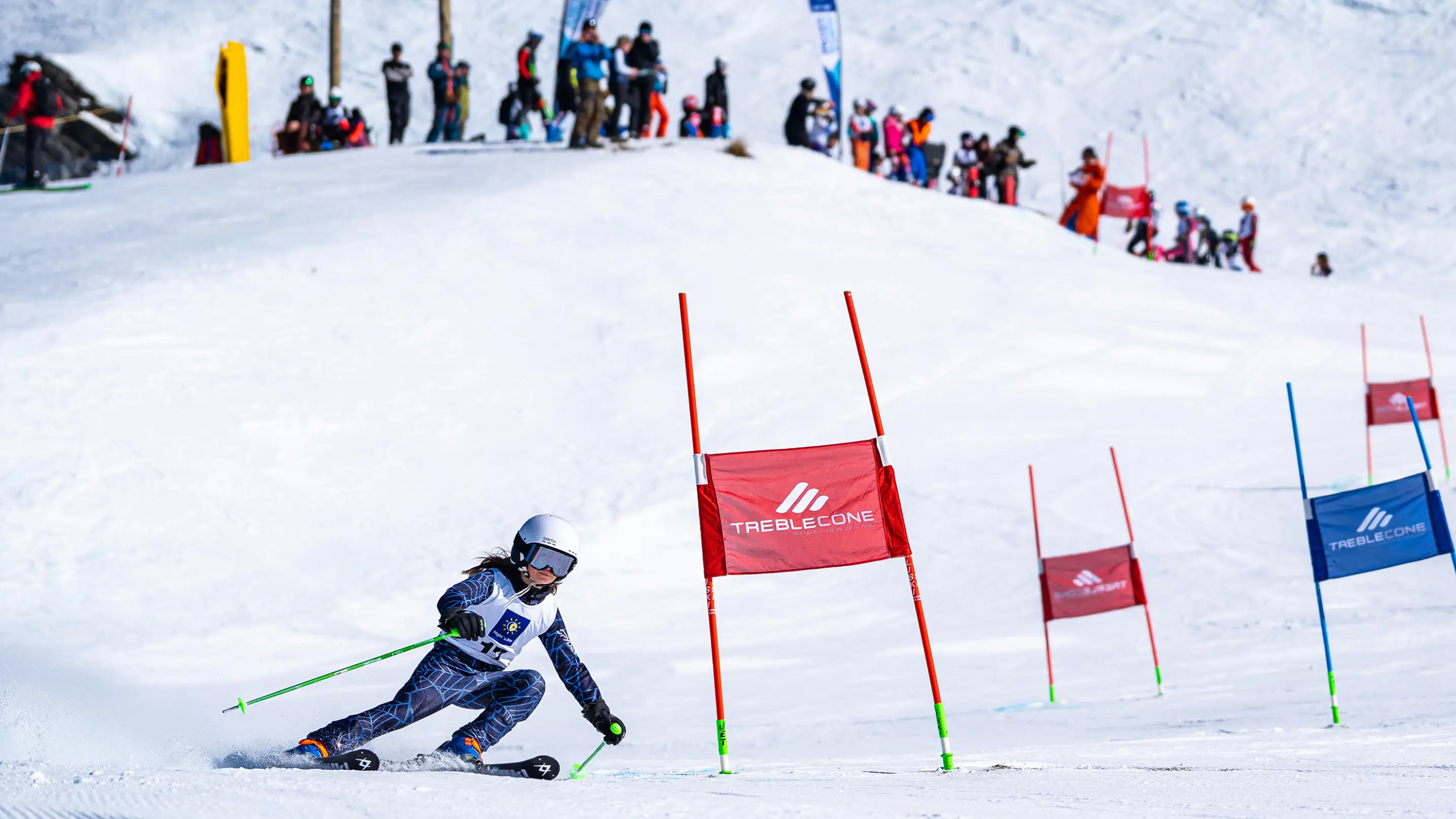 A young female ski racer carves through red Treble Cone branded gates during a giant slalom race, with a crowd of spectators watching from the snowy hillside above.