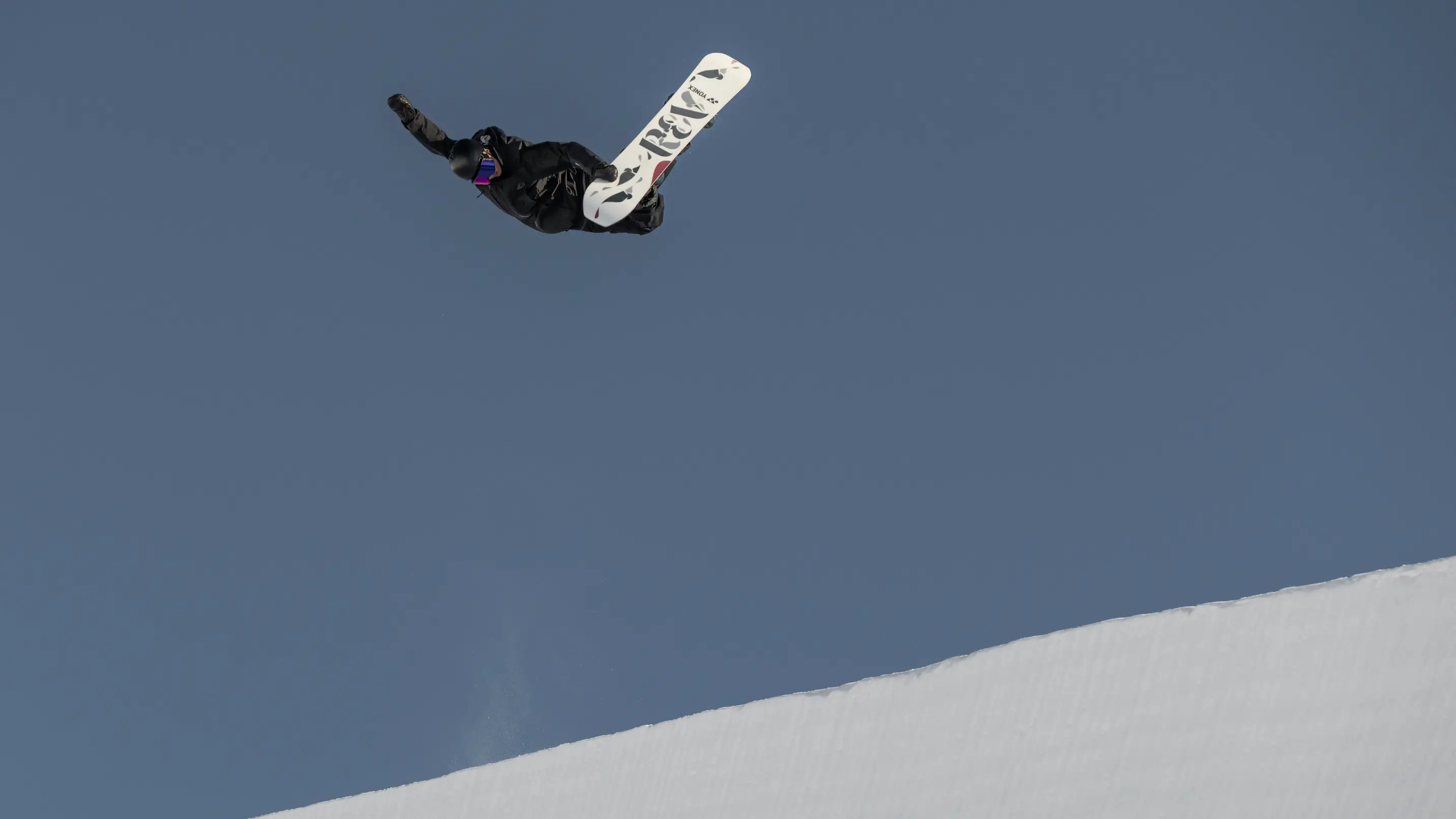 A snowboarder performs an inverted trick high above the halfpipe lip against a clear blue sky.