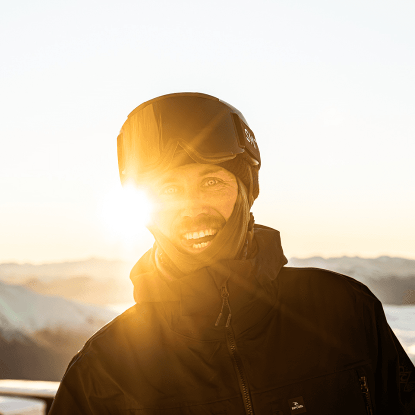 Headshot of a smiling Stef Zeestaten, NZ skier, standing outside on a snowy mountain wearing a black jacket, buff and ski goggles.