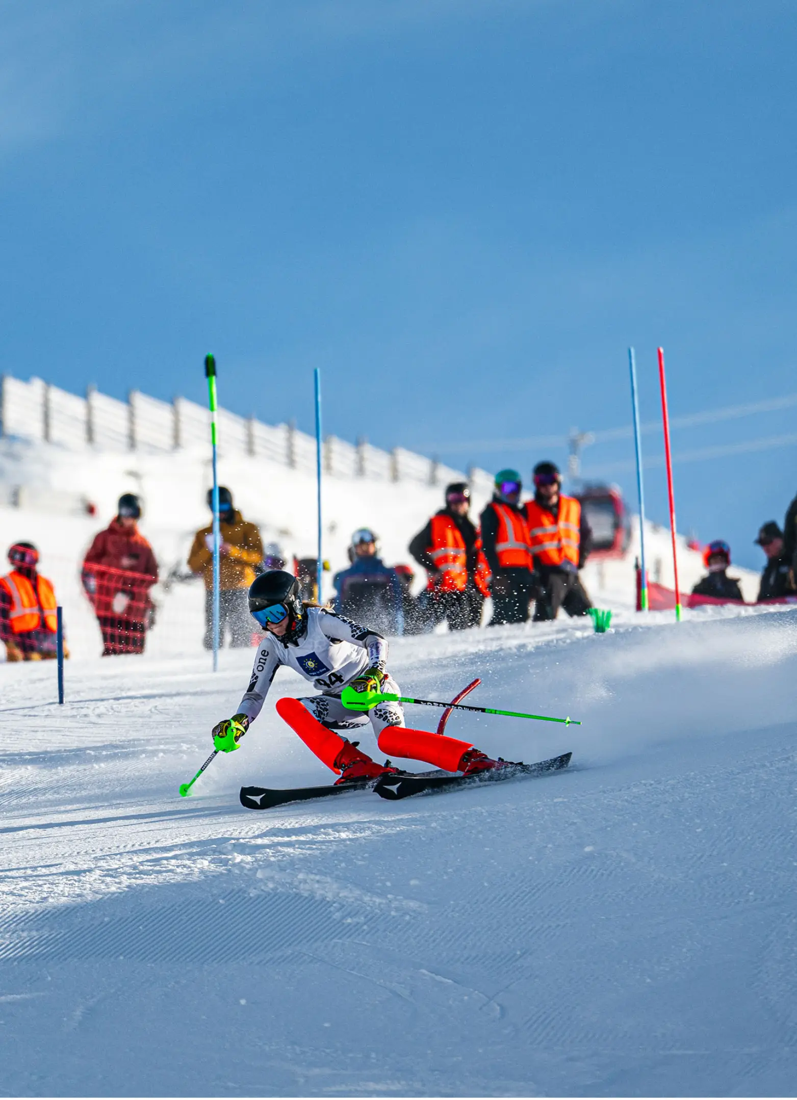 A female slalom ski racer in a white and red suit carves aggressively through a gate, with orange-vested officials and spectators watching from the sideline.