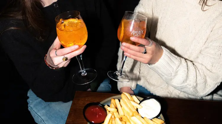 Two friends sitting side by side have an aperol spritz each and a plate of fries to share.