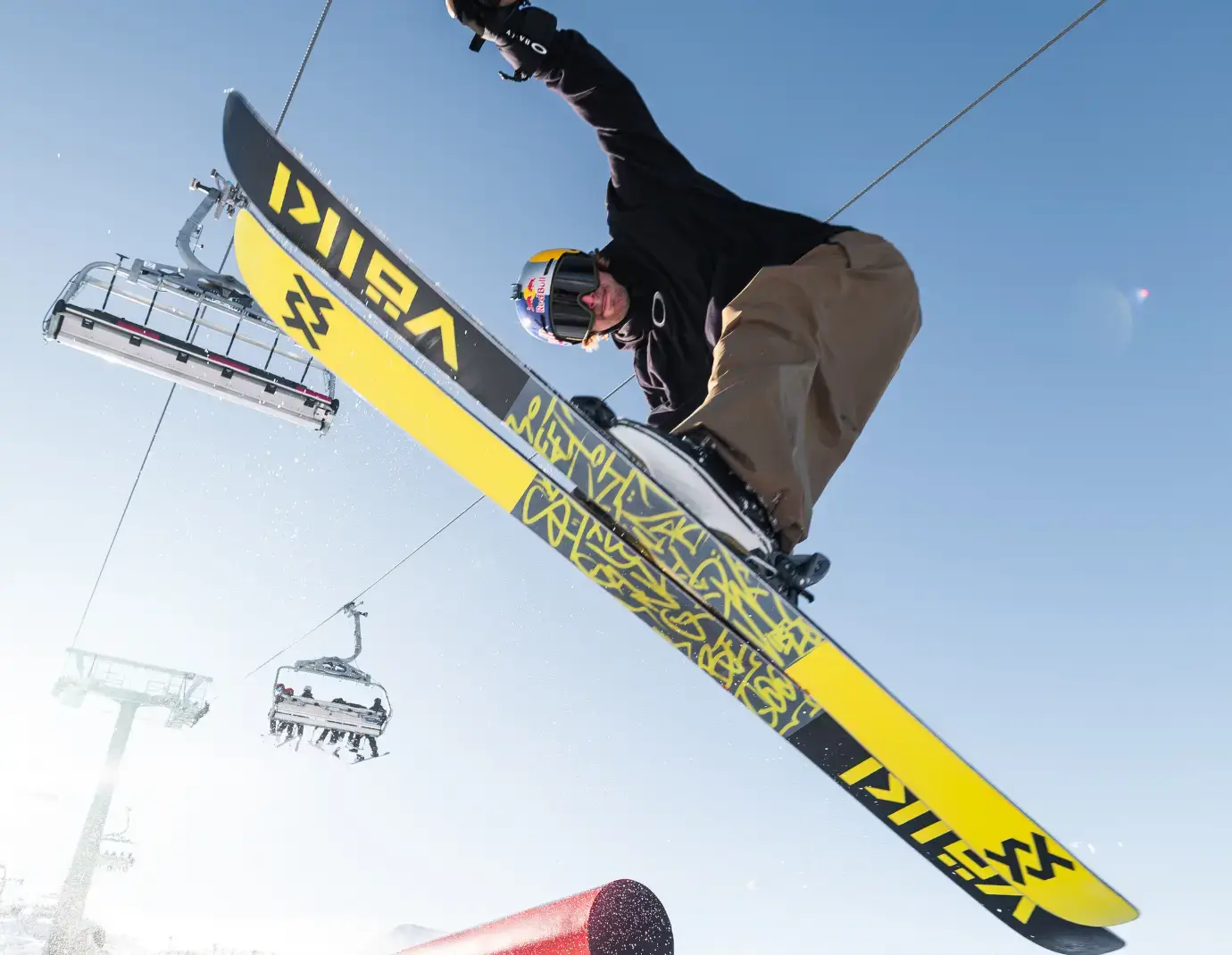 A skier performs an aerial trick under a clear blue sky, with a ski lift and mountain backdrop visible in the background.