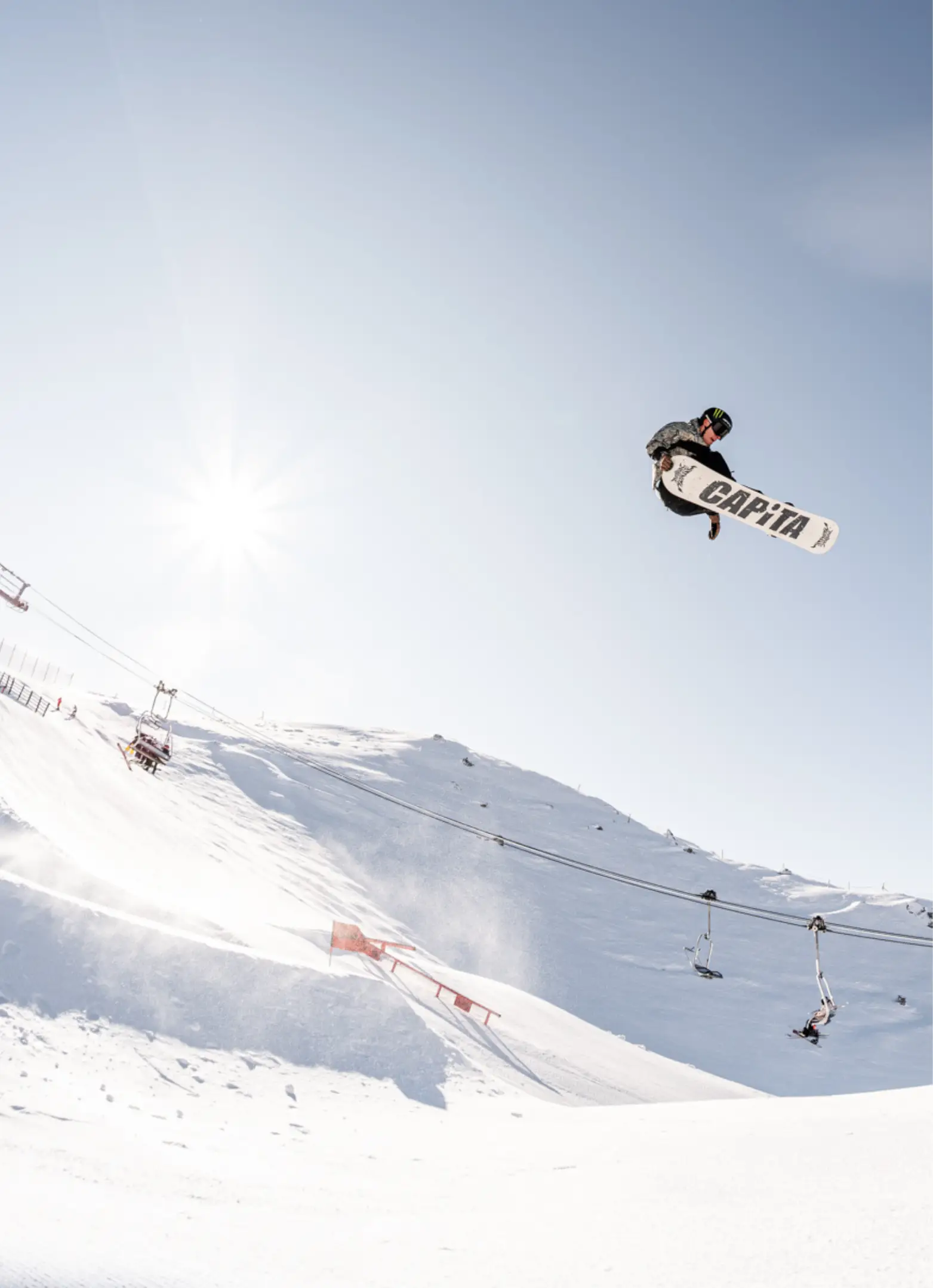 A snowboarder on a CAPiTA board soars above a large kicker in the terrain park with chairlifts and a sunny alpine landscape below.