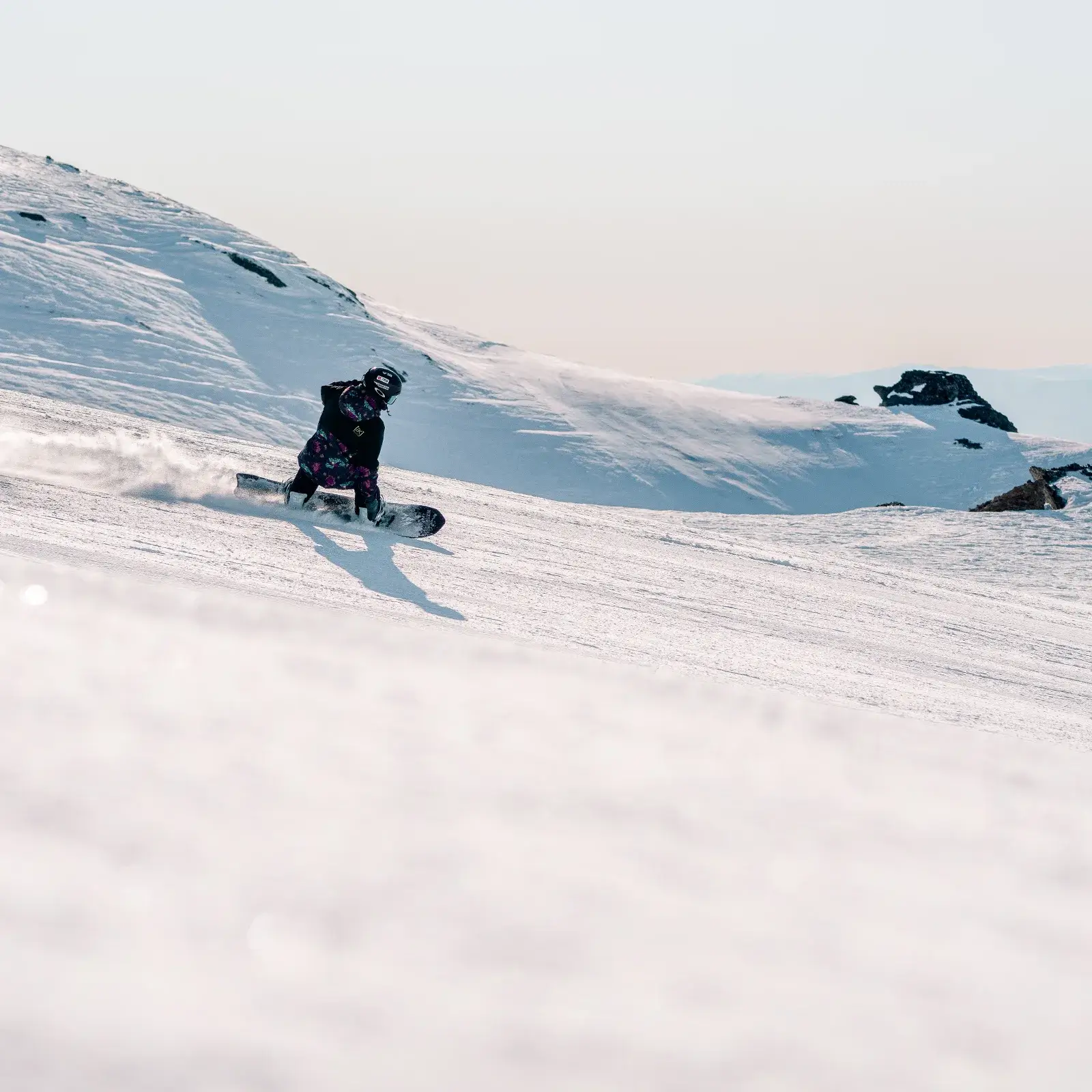 Snowboarder carving on a wide, snowy slope with a clear sky and rugged mountain peaks in the background.