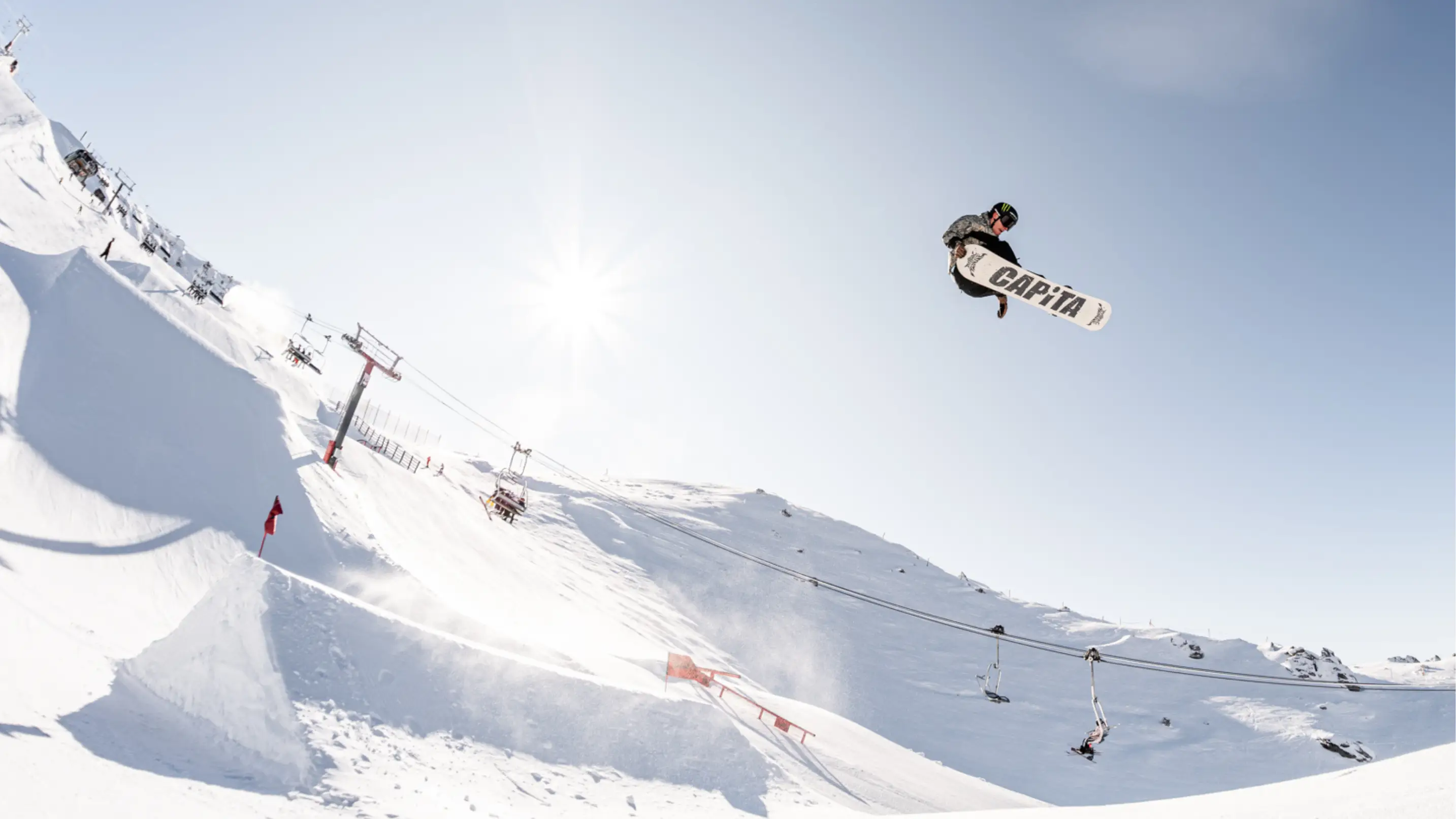 A snowboarder on a CAPiTA board soars above a large kicker in the terrain park with chairlifts and a sunny alpine landscape below.