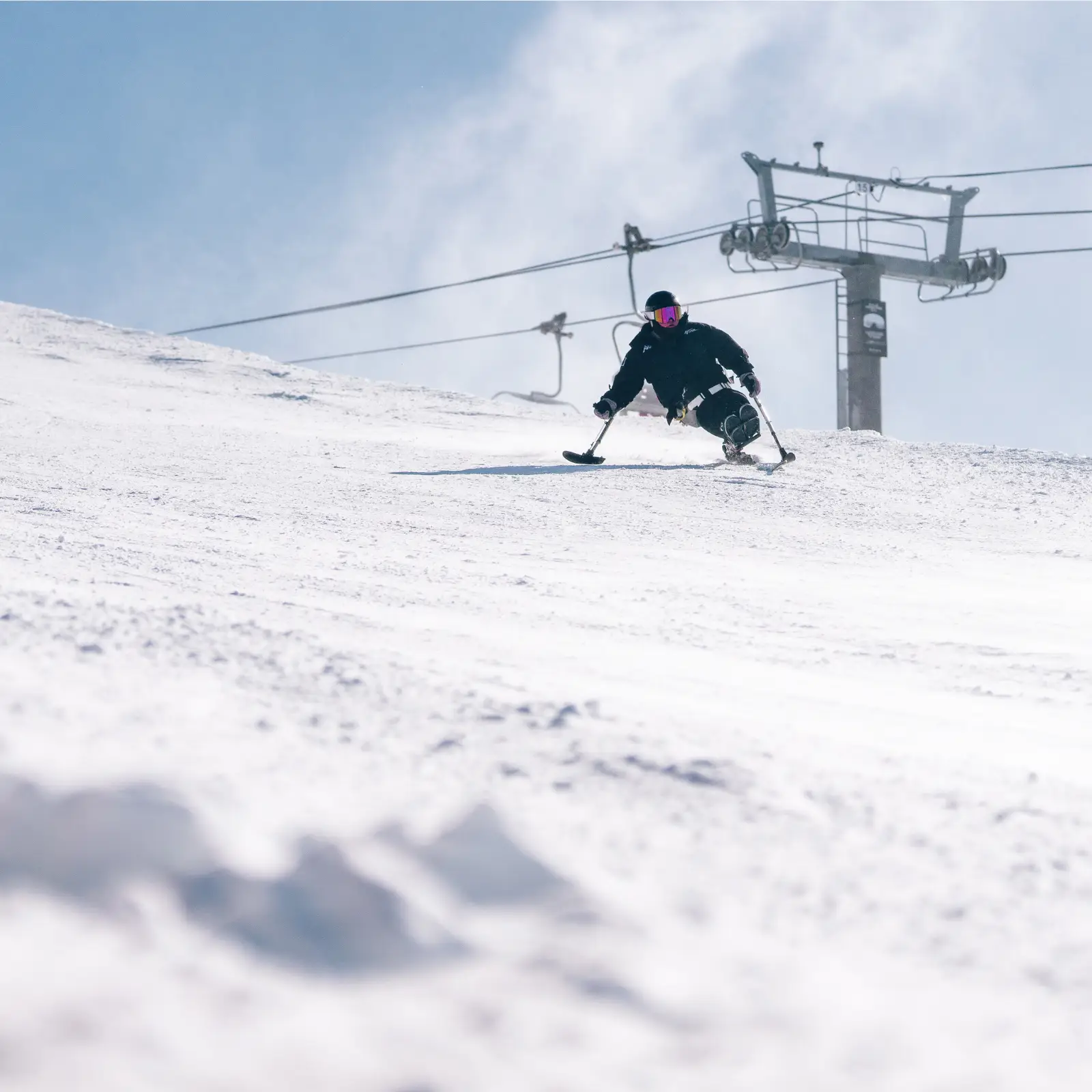 Adaptive sit-skier carving down a snowy slope with a chairlift in the background on a bright winter day.