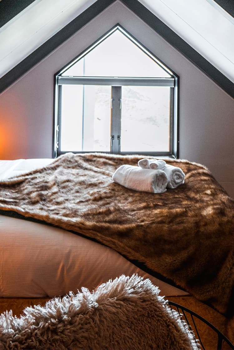 A cozy attic bedroom featuring a sloped ceiling with exposed beams, a comfortable bed draped with a soft fur blanket, and warm lighting, creating an inviting atmosphere at the Cardrona Apartments.