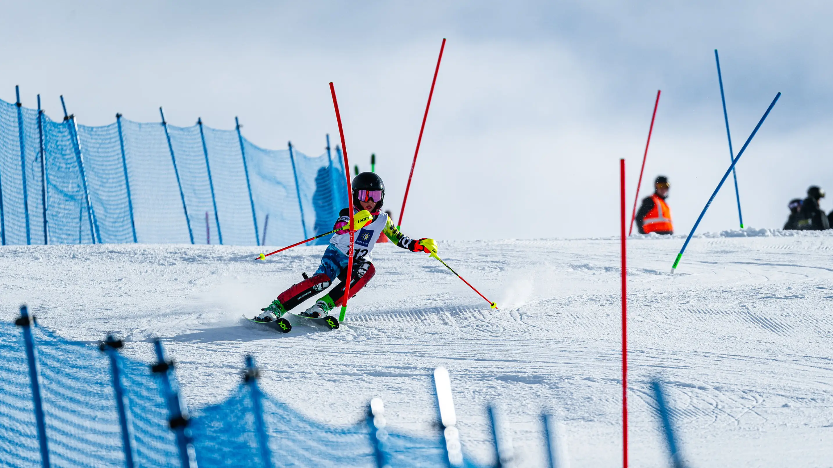 A young junior alpine ski racer in a colourful race suit navigating a slalom course, carving past red and blue gates on a groomed snow slope.