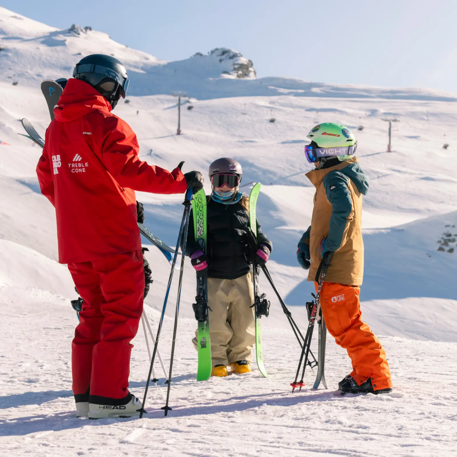 Ski instructor in a red suit talking with two children holding skis on a snowy mountain slope.