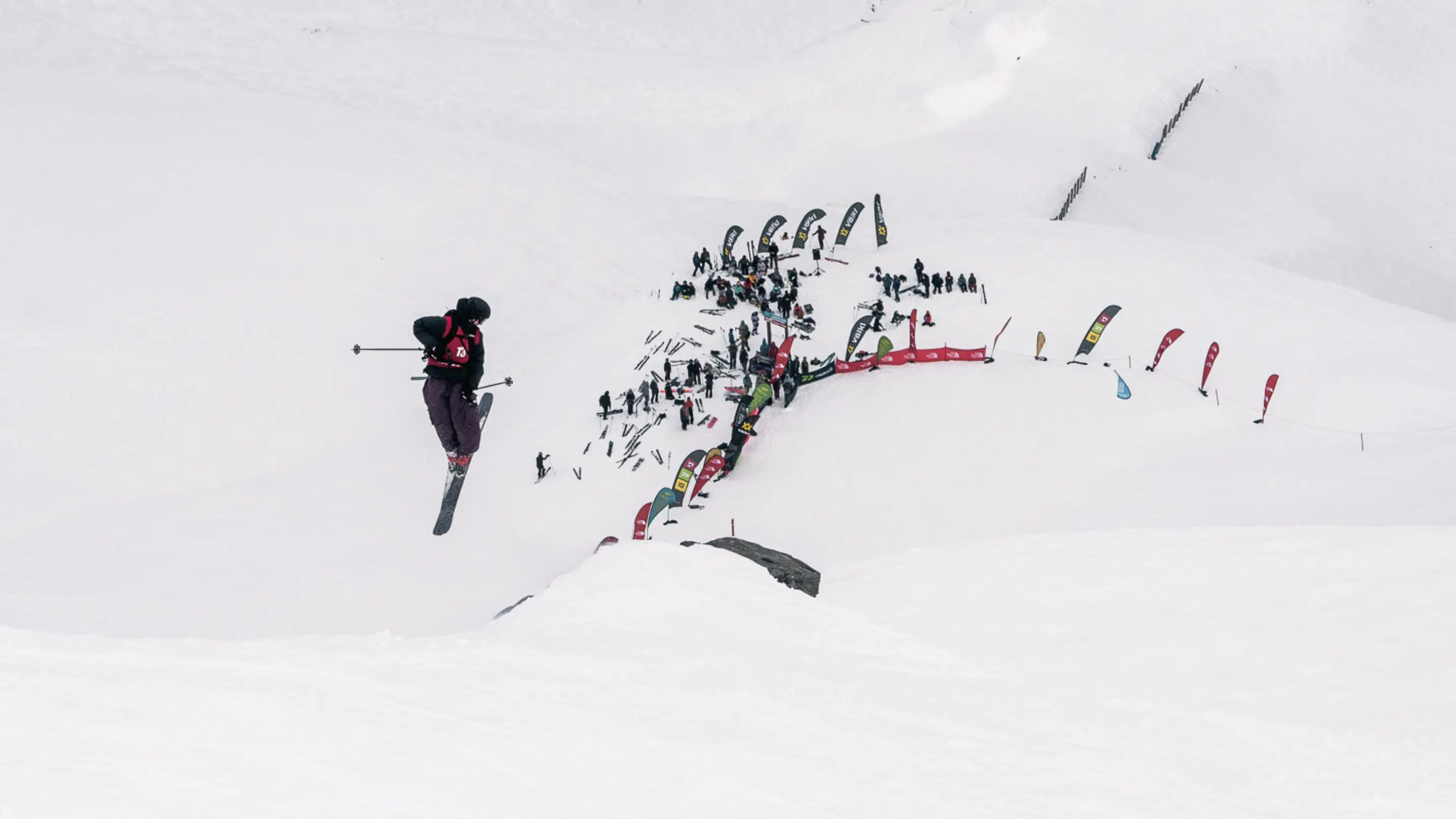 A freeskier launching off a kicker high above a crowd of spectators and sponsor flags on a snowy slope.