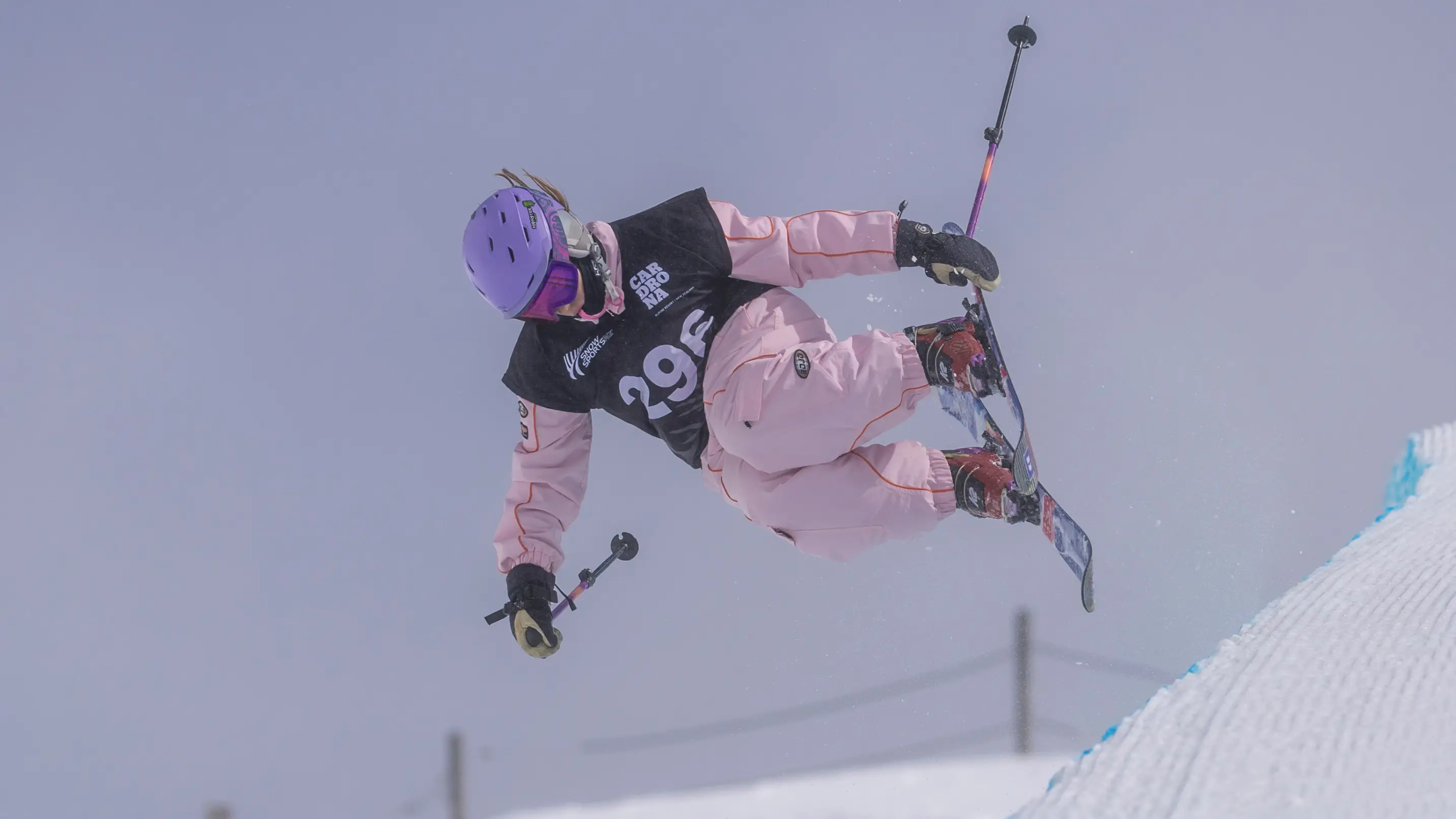 A freeskier wearing bib 29, in a pink outfit and purple helmet, grabs their ski mid-air above a halfpipe at Cardrona, with spectators watching below.