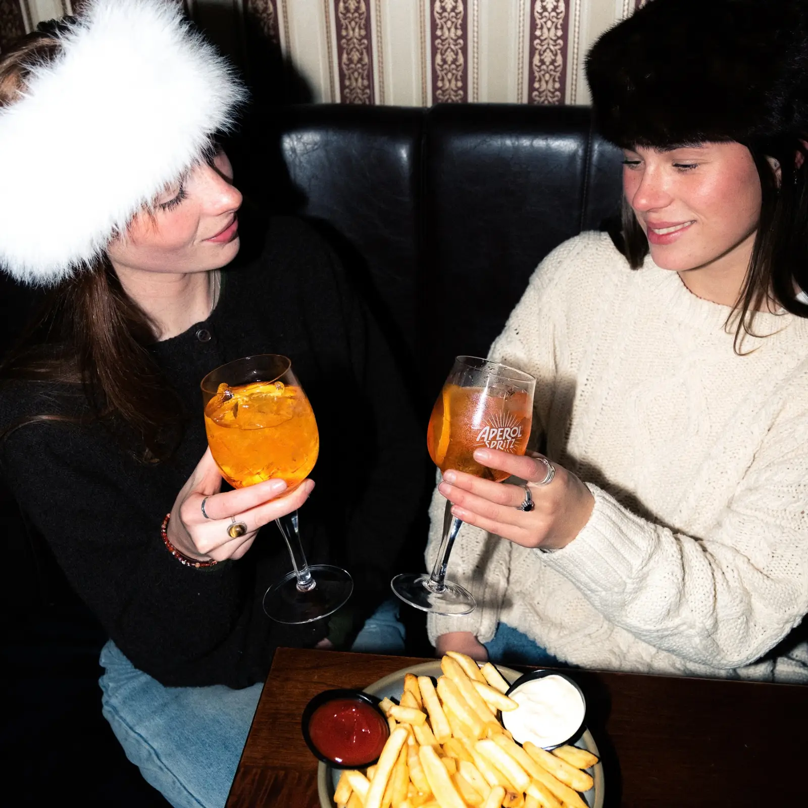 Two friends sitting side by side have an aperol spritz each and a plate of fries to share.