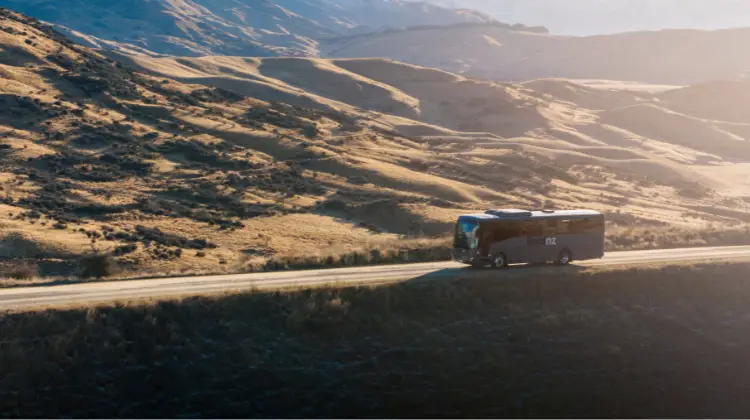 A tour bus travels along a winding road through a golden, mountainous landscape.