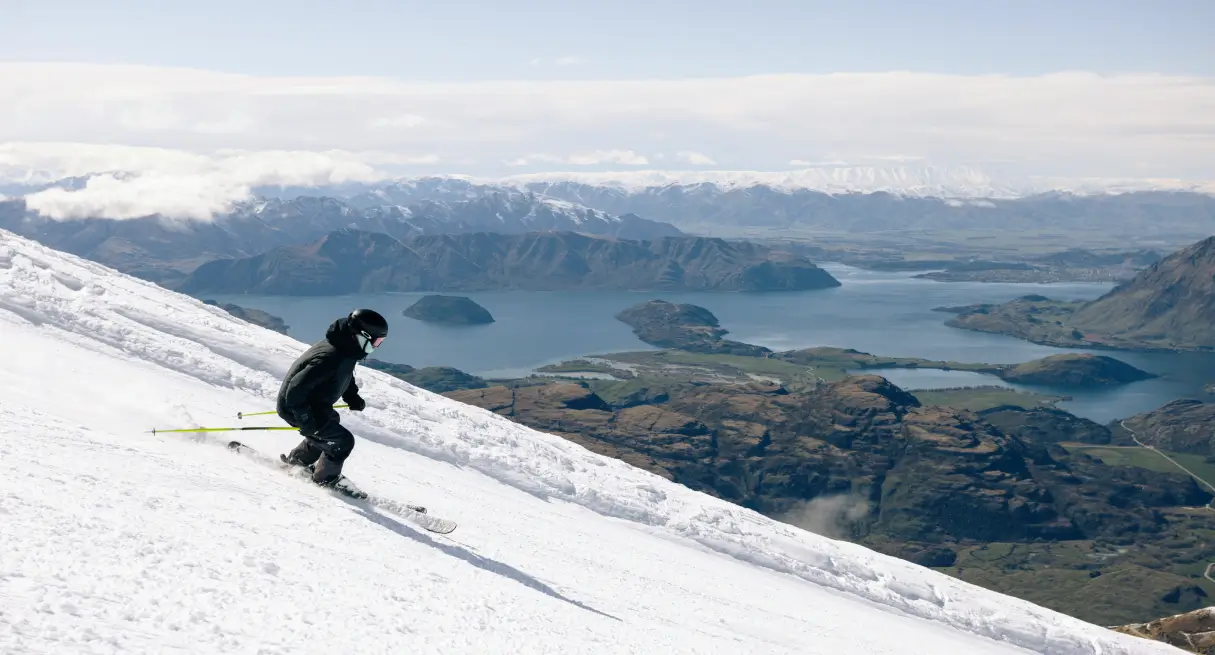 Skier carving down a snowy slope with stunning views of Lake Wānaka and surrounding mountains in the background at Treble Cone, New Zealand.
