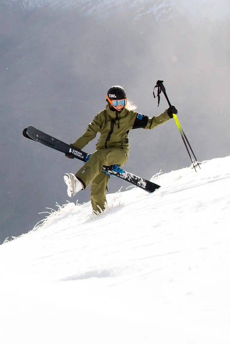 A person in ski gear joyfully hikes up a snowy mountain slope carrying skis and poles, with a dramatic alpine valley and braided river in the background.