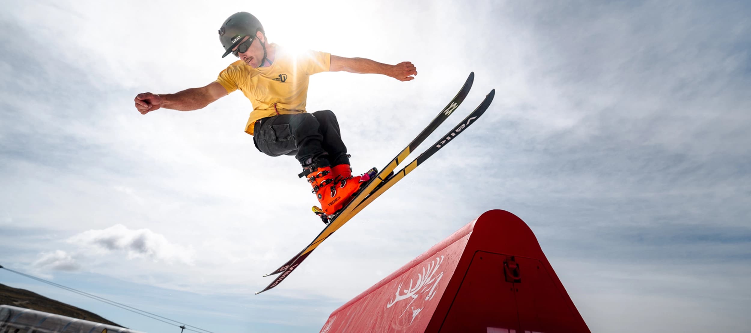 Skier in a helmet and yellow shirt launches into the air above a red terrain-park feature, with bright sun and blue sky behind.