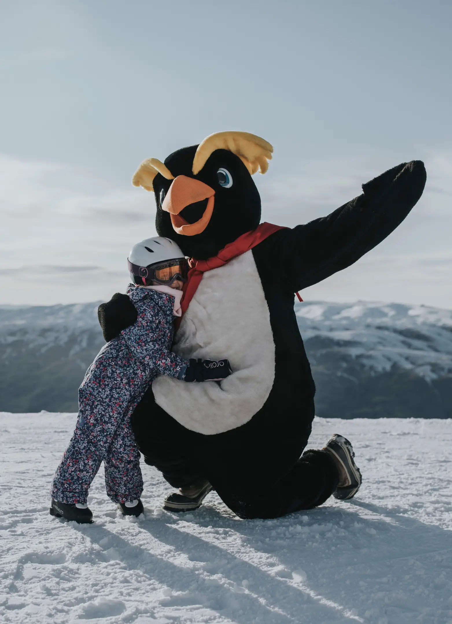 A penguin mascot poses hugging a child, dressed in full ski gear, on a snowy mountain.
