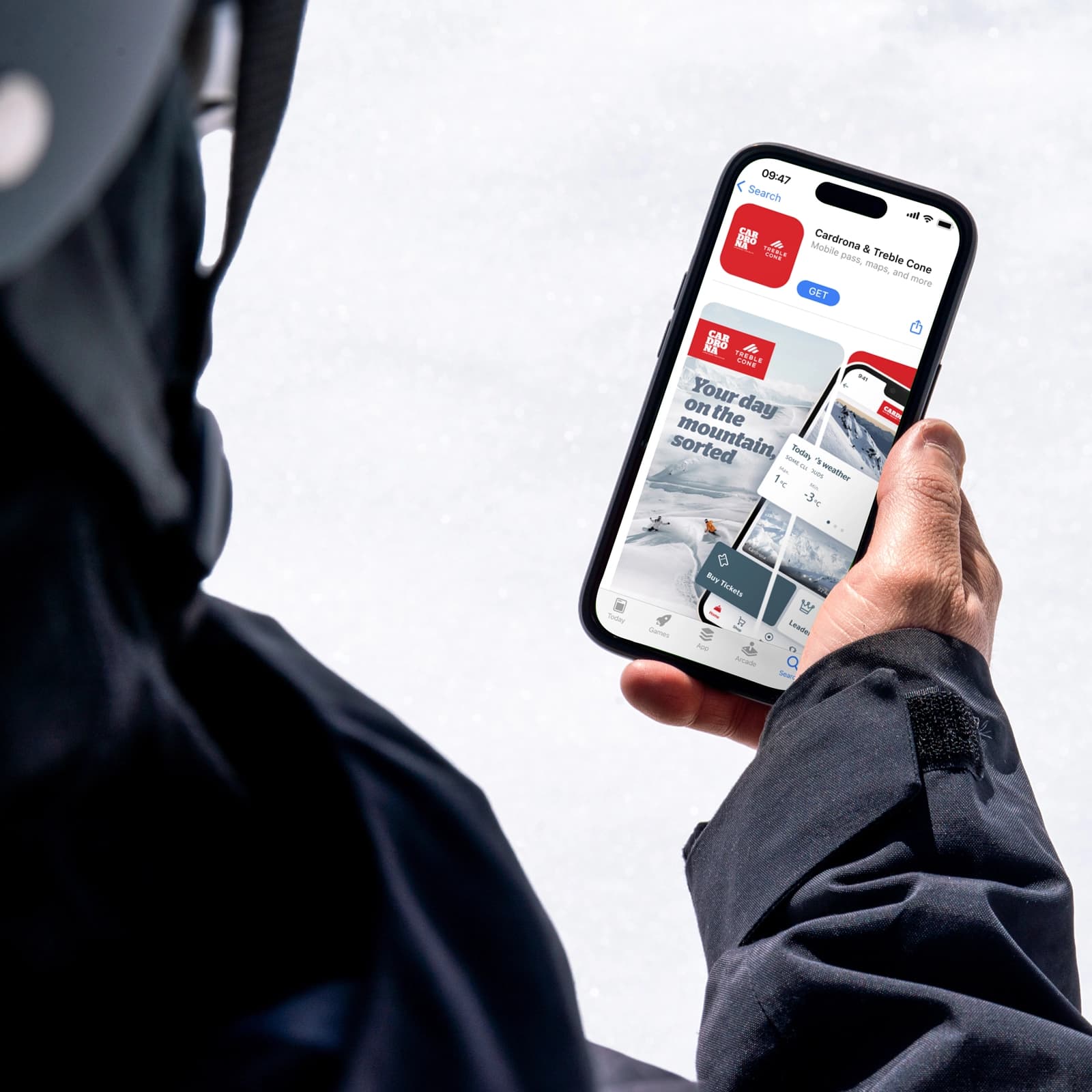 A skier in a helmet and jacket holding a smartphone showing the Cardrona & Treble Cone app listing on the Apple App Store, with a snowy slope in the background.
