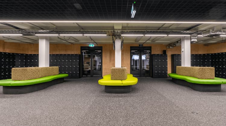 A modern locker room with black lockers lining the walls, green and tan curved bench seating in the center, and industrial-style ceilings with exposed piping and lighting.
